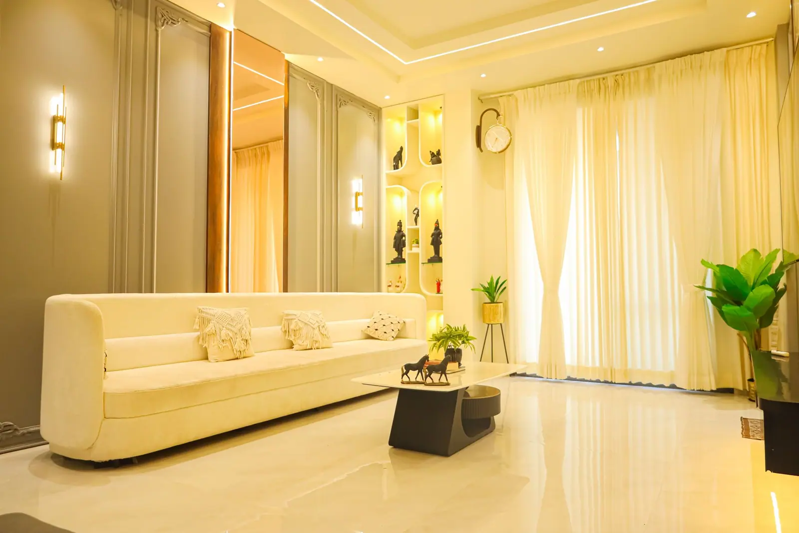 Living room — a curved cream sofa against sheer drapes and a lit display wall.